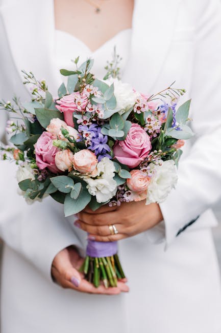 A person in a white outfit holding a floral bouquet with both hands, featuring pink roses, pale pink spray roses, white and light purple lisianthus, small blue delphiniums, and sprigs of eucalyptus wi