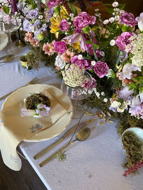 A floral arrangement featuring a variety of fresh flowers, including pink and purple peonies, white and lavender roses, delicate daisies, and clusters of small white blossoms, arranged in a tall, clea