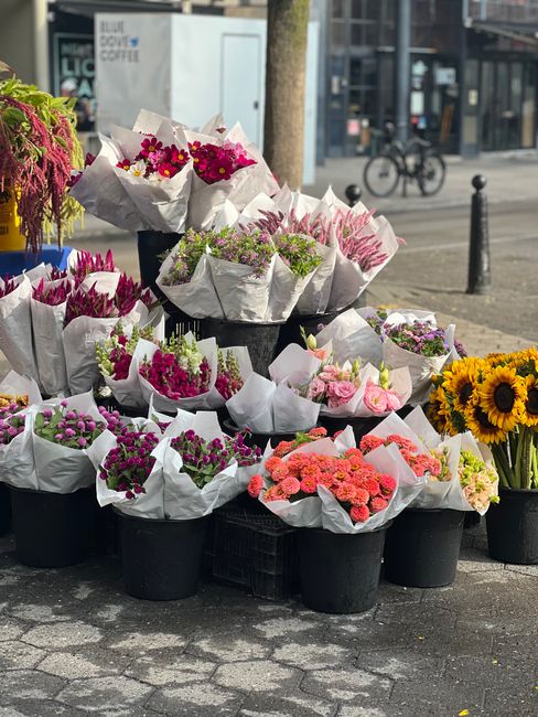 A display of various fresh flower bouquets arranged in black pots and black crates outdoors on a paved sidewalk, with some bouquets wrapped in white tissue paper featuring pink, purple, and yellow flo