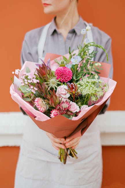 A person in a light grey shirt and white apron holds a large bouquet of fresh flowers wrapped in pink and brown paper. The bouquet features vibrant pink, white, and green blossoms, including chrysanth